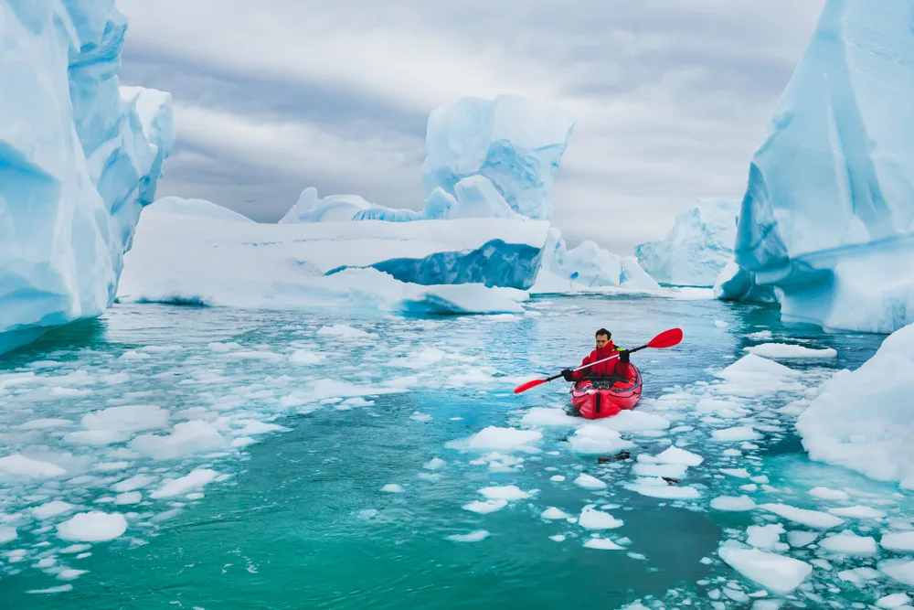 Man in a red kayak paddling through the giant icebergs during the overall cheapest time to visit Antarctica