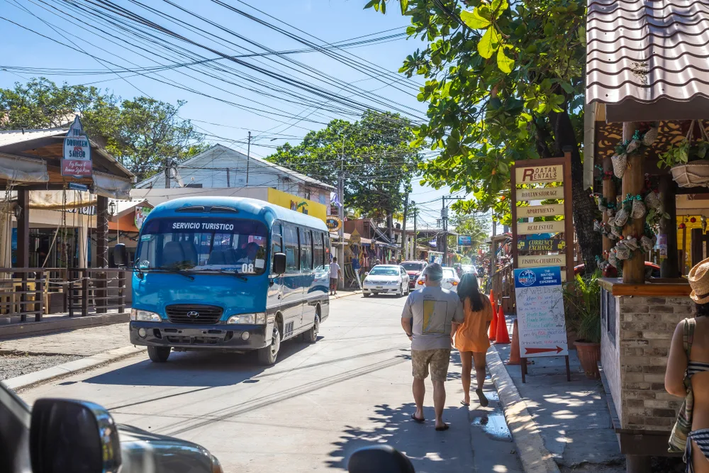 Inland photo of a market in Roatan pictured during the best time to visit with a colorful blue bus next to people walking by the market
