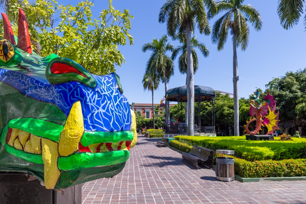 Blue, green, and yellow dragon sitting in the foreground of a city park with benches all around pictured during the best time to visit Mazatlan