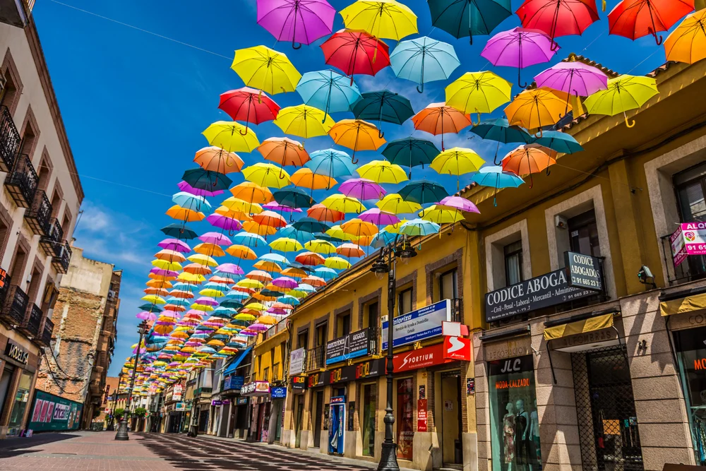 Photo of brightly-colored umbrellas hanging from strings below a deep blue sky between old colonial-style buildings in Madrid, as seen during the best time to visit