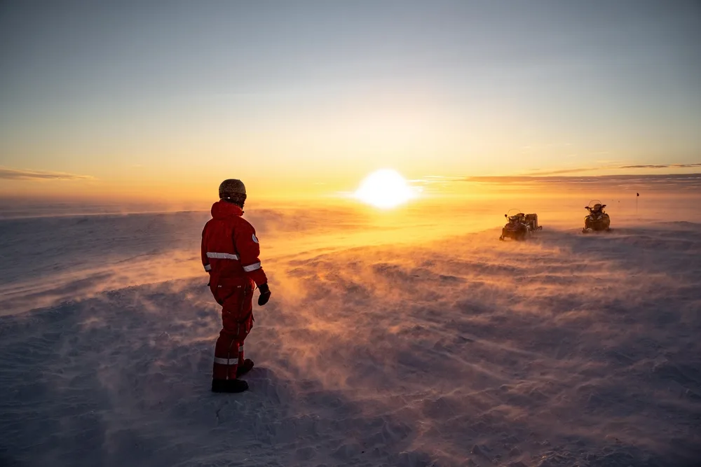 Pictured during the least busy time to visit Antarctica, a man by a snowmobile stands and watches the sun set over the eternal snowy horizon