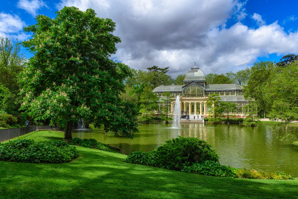 View of the gorgeous Crystal Palace (Palacio de Cristal) as seen from the shoreline of the lake with a fountain in front during the overall best time to visit Madrid, the spring