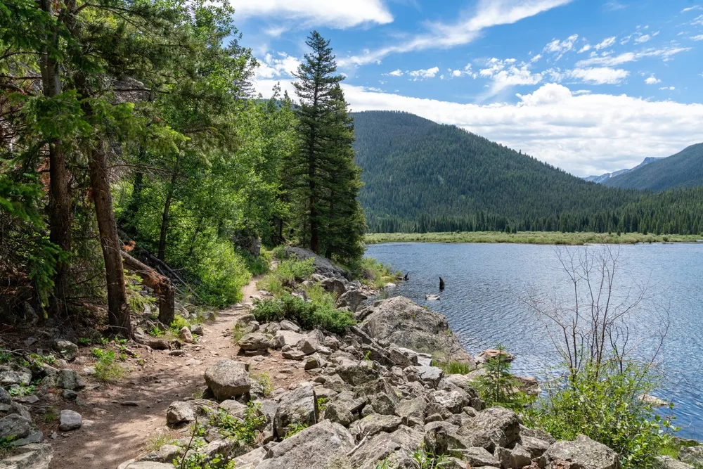 Hiking trail around a deep blue lake pictured during the overall best time to visit Rocky Mountain National Park, the spring or summer