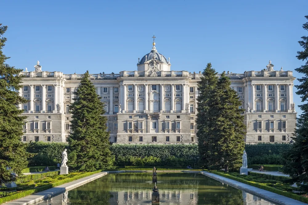 View of the stately Royal Palace of Madrid pictured from the front with blue skies overhead and a reflecting pool in the foreground, surrounded by statues