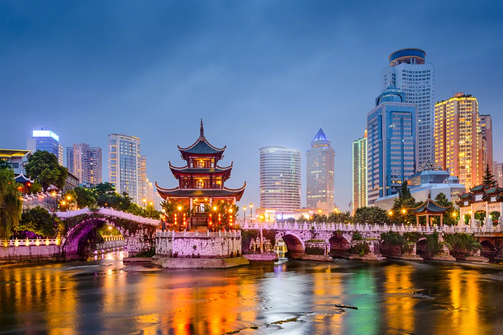Photo of the Guiyang China skyline with the Jiaxiu Pavilion pictured in the foreground, all lit up at night, with reflections of the tall buildings on the water