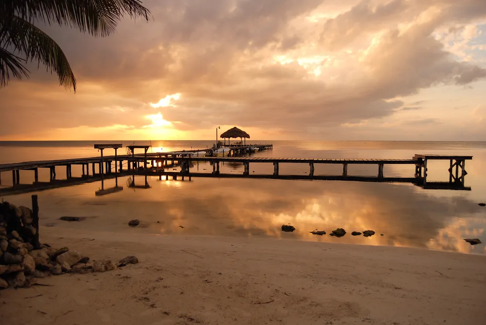 Dock pictured below a setting sun with still water and clouds overhead during the winter, the cheapest time to visit Belize
