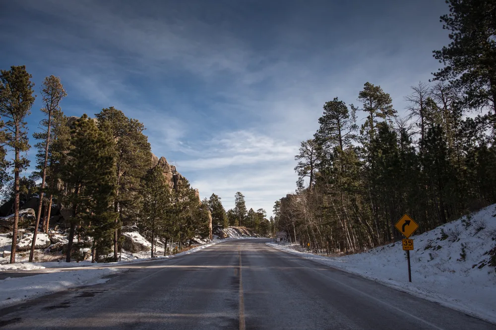 Pictured during the winter, the overall worst time to visit Mount Rushmore, a snow road is pictured running down the middle of a snow-capped valley with green trees lining either side
