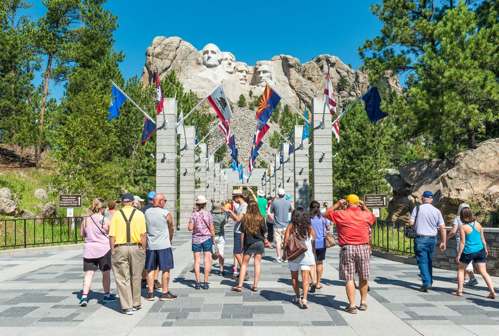 Photo of a small crowd of people standing in front of Mount Rushmore an the many flags that line the walkway to the edge of the park seen on a clear blue-sky day