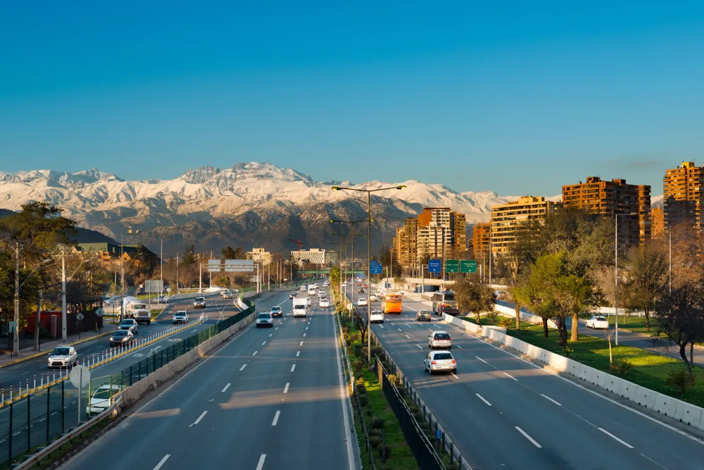 Expressway with snow-capped mountains in the background pictured during the overall worst time to visit Santiago, Chile, with a highway running down the middle of the scene