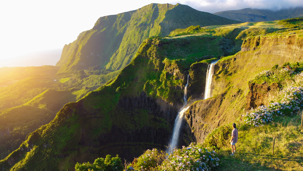 Lush green waterfall landscape on Flores Island, pictured during the best time to visit the Azores, with water falling hundreds of feet and a lone tourist looking over the scene
