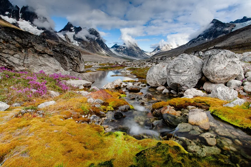 Glacier stream surrounded by tall mountains and brown vegetation pictured during the cheapest time to visit Greenland