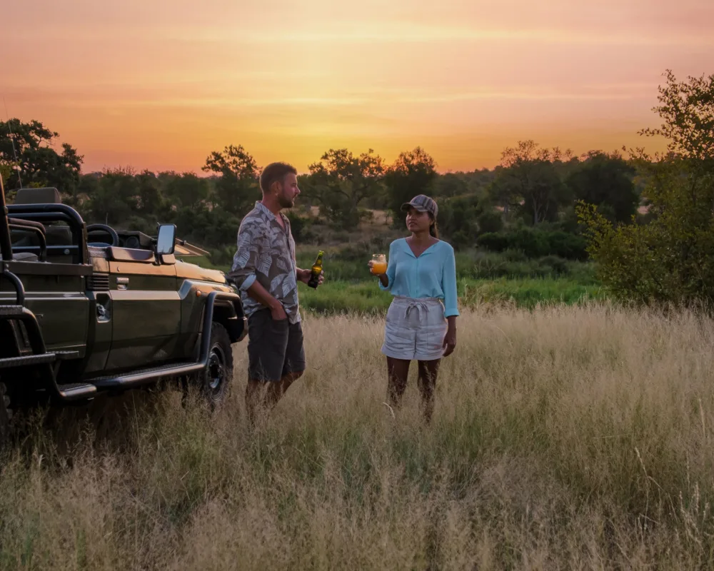 Asian woman and a man standing in a field during the best time to visit Kruger National Park with a nice orange sky setting over the field behind them
