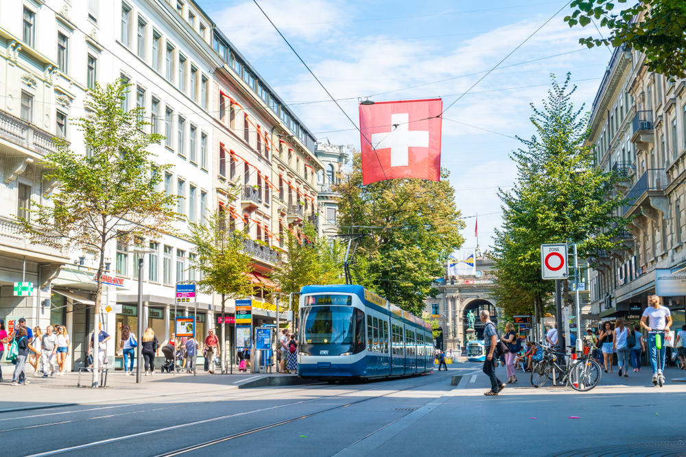 Photo of the middle of town with historical buildings and people surrounding a tram pictured under a blue sky during the best time to visit Zurich