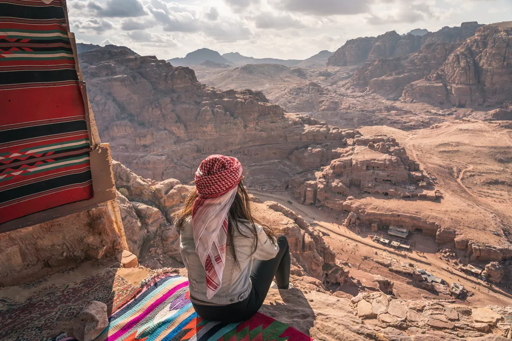 Woman sitting on a colorful Arab carpet looking at the Petra ruins in Jordan for a guide to whether or not the country is safe to visit