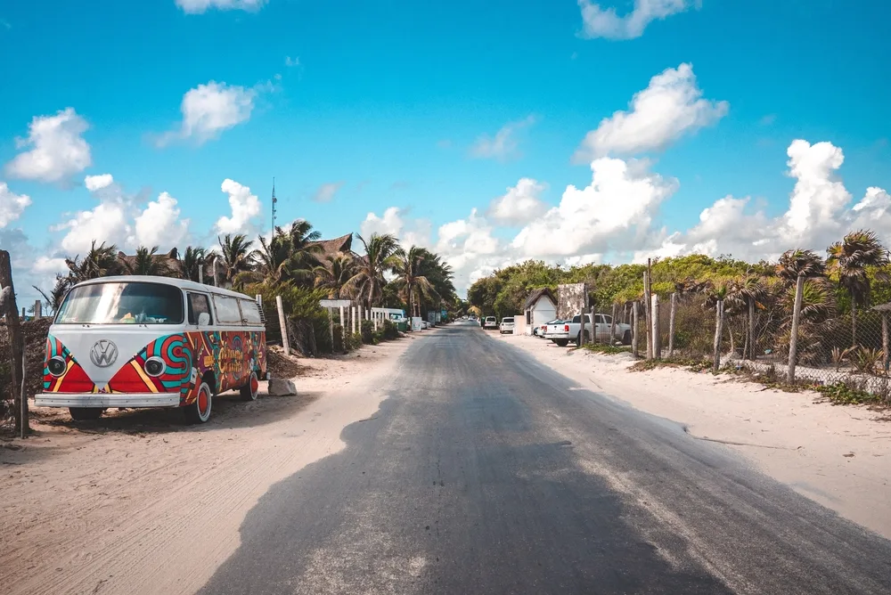 Photo of a VW bug sitting next to an asphalt road with sand on either side of it pictured during the cheapest time to visit Tulum