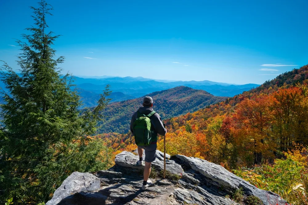 Man relaxing at a scenic overlook in the fall, the overall best time to visit Asheville NC, with a walking stick in his hand and his leg up on a rock