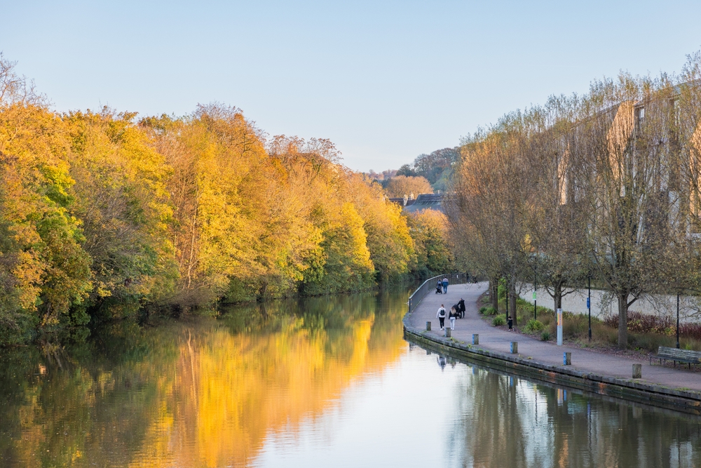 Postcard-worthy view of golden trees and still water with people walking along the river pictured during the cheapest time to visit the United Kingdom, in autumn