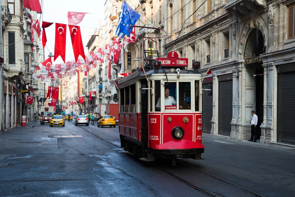 To help answer the question "Is Istanbul Safe to Visit" a photo of a red trolley making its way through the historic old buildings is shown in an otherwise tranquil scene
