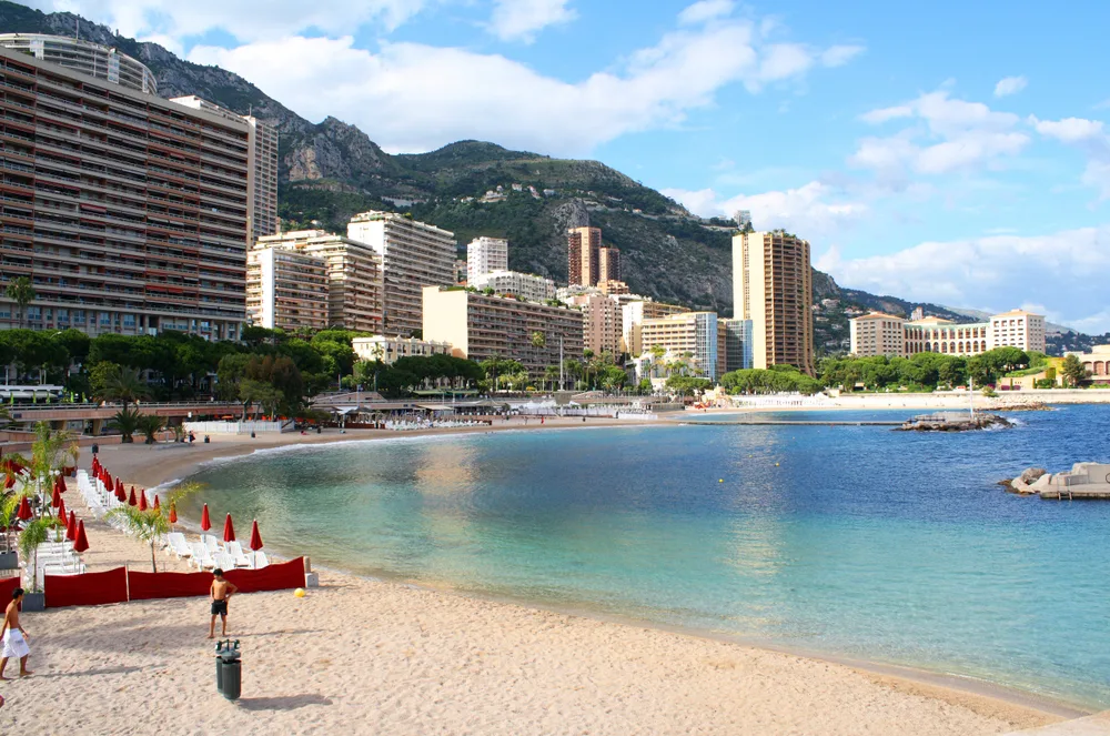 Larvotto Beach, shaped like a crescent with beach chairs lined up and the skyline of Monte-Carlo with blue lightly cloudy skies during the best time to visit Monaco