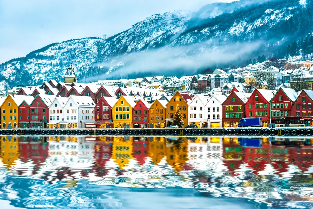 View of Bruges, Bergen with snowy mountains and colorful buildings reflecting into the water during the cheapest time to visit Norway in winter