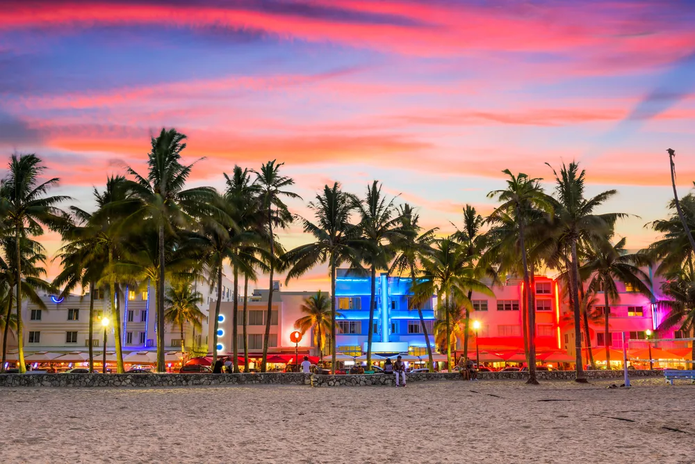 Colorful sunset skies over Ocean Drive lined with palm trees and neon-lit buildings with the beach visible in the foreground during the best time to visit Miami